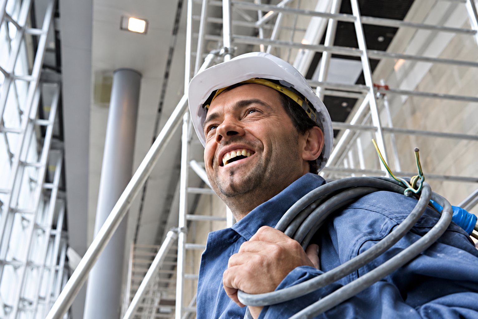 Ouvrier souriant en casque blanc portant des câbles, devant un échafaudage