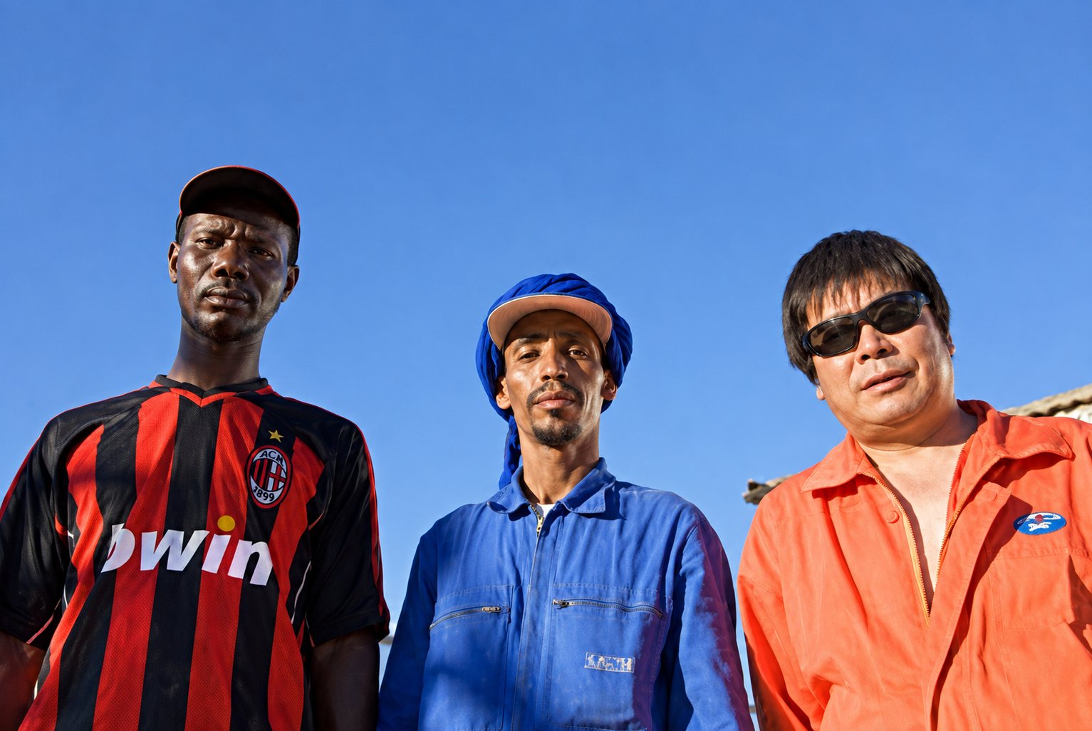 Trois ouvriers en maillot rayé, bleu de travail et combinaison orange, sous un ciel bleu