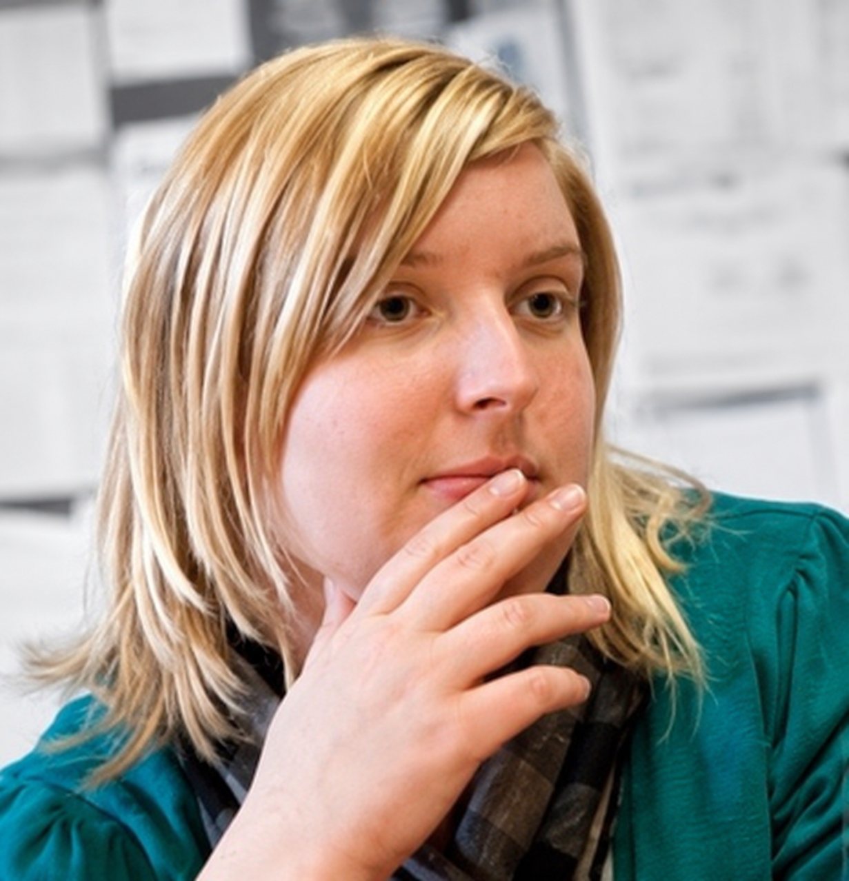 Jeune femme blonde pensive, la main devant la bouche, veste verte et écharpe, devant un mur de documents