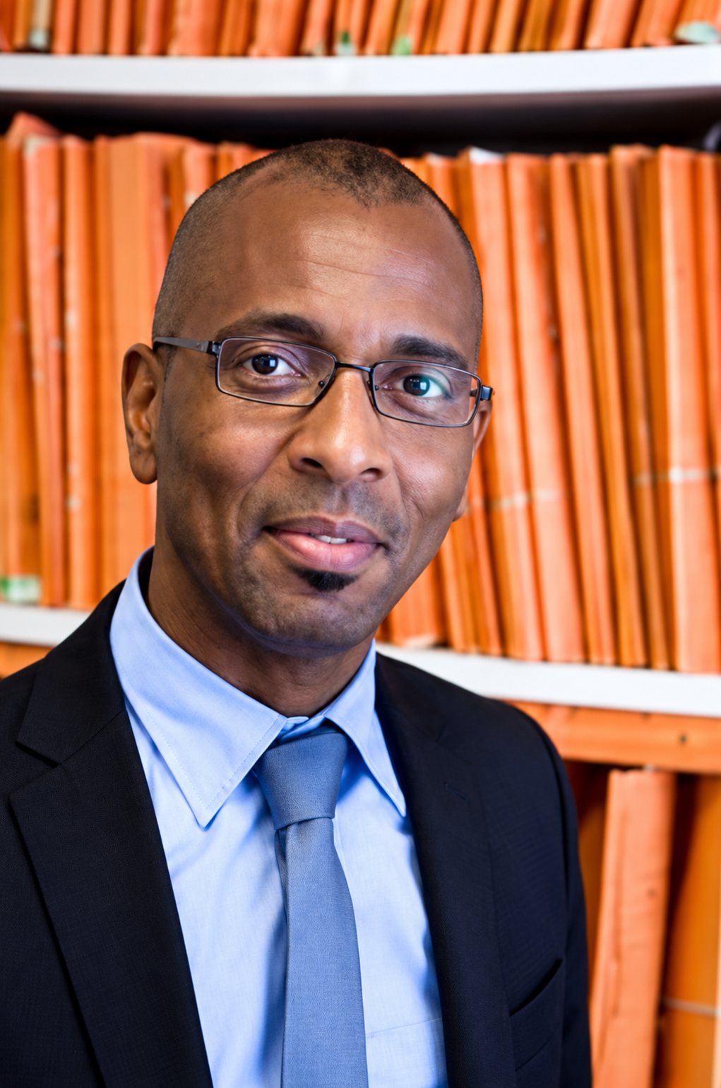 Homme à lunettes en costume sombre devant des classeurs orange
